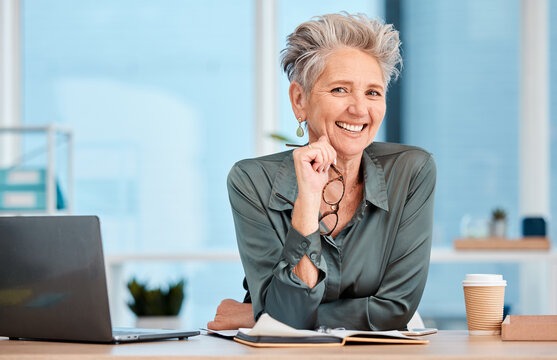 Smiling older woman with short gray hair sits at a desk, holding glasses and surrounded by a laptop, notebook, and coffee cup, exuding confidence.